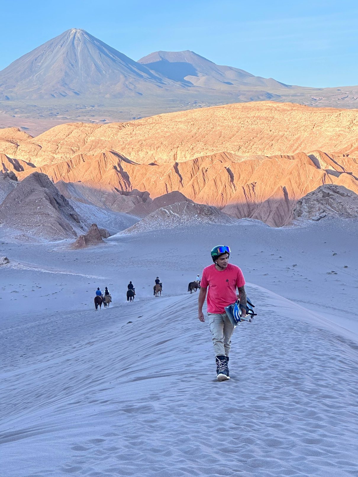 Instructor caminando por una duna del Valle de la Luna, San Pedro de Atacama, con el volcán Licancabur al fondo, tabla de sandboard bajo el brazo