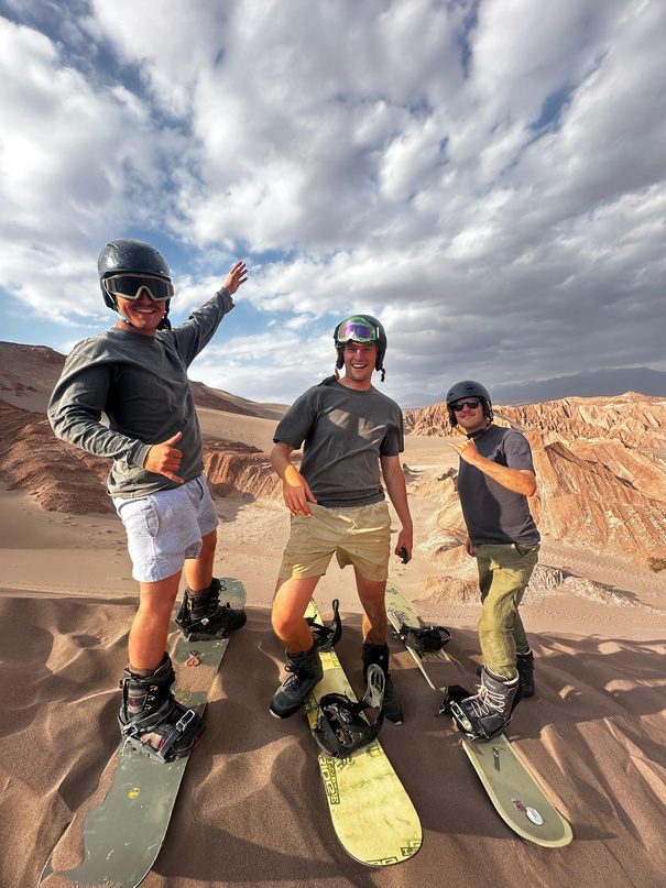 Tres personas sentadas en la arena con sus tablas de sandboard, cielo nublado